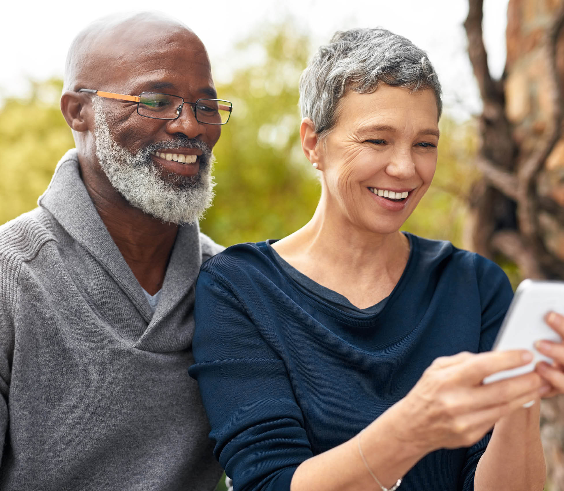 Interracial senior couple, phone and smile in park, garden or nature for love or care together. Happy man, woman and partner for retirement, relax and support for marriage and vacation for pensioner.
